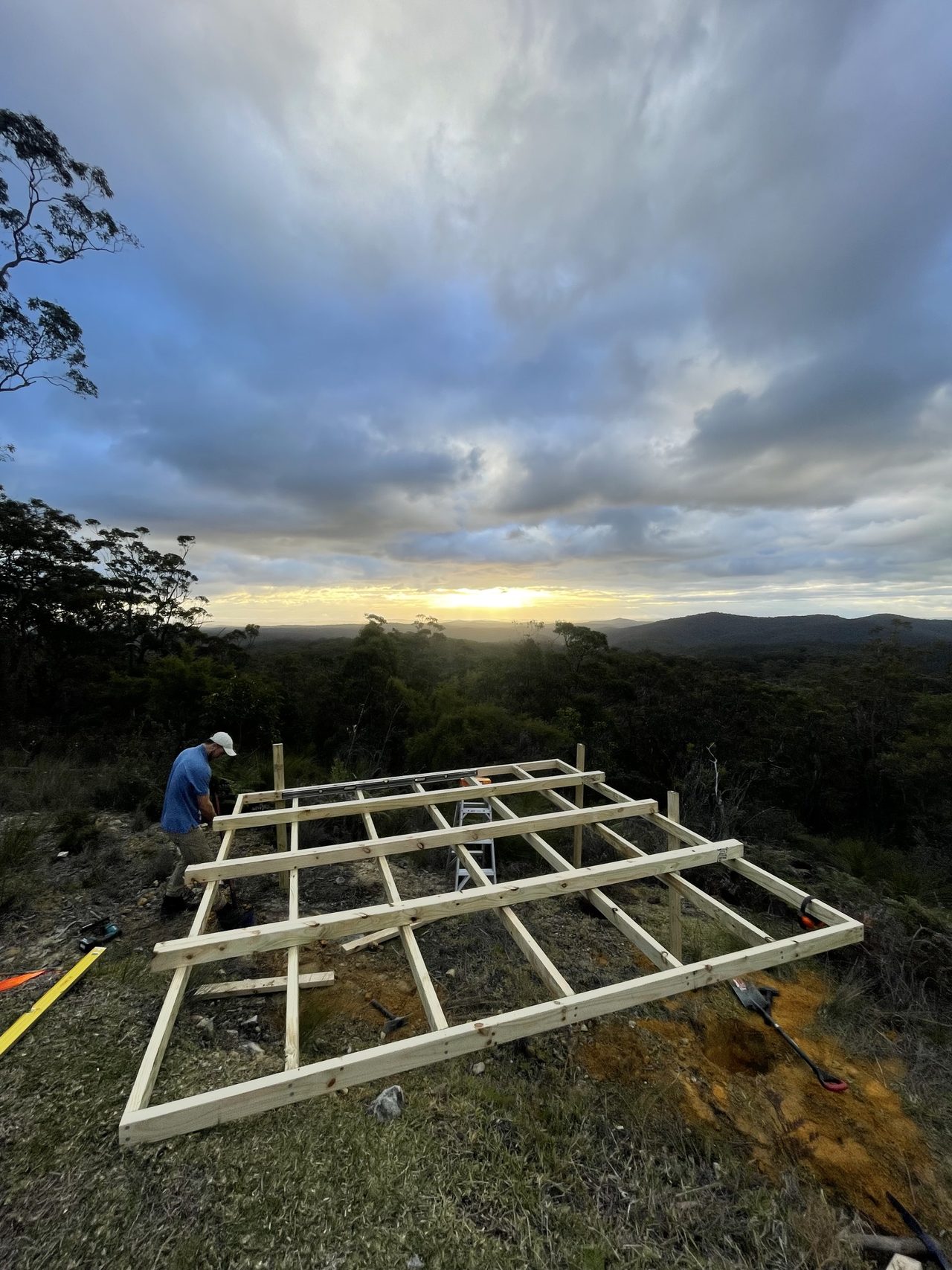 A timber platform being framed on a cleared bush ridge at sunset, with a person working on the frame and ranges in the distance.