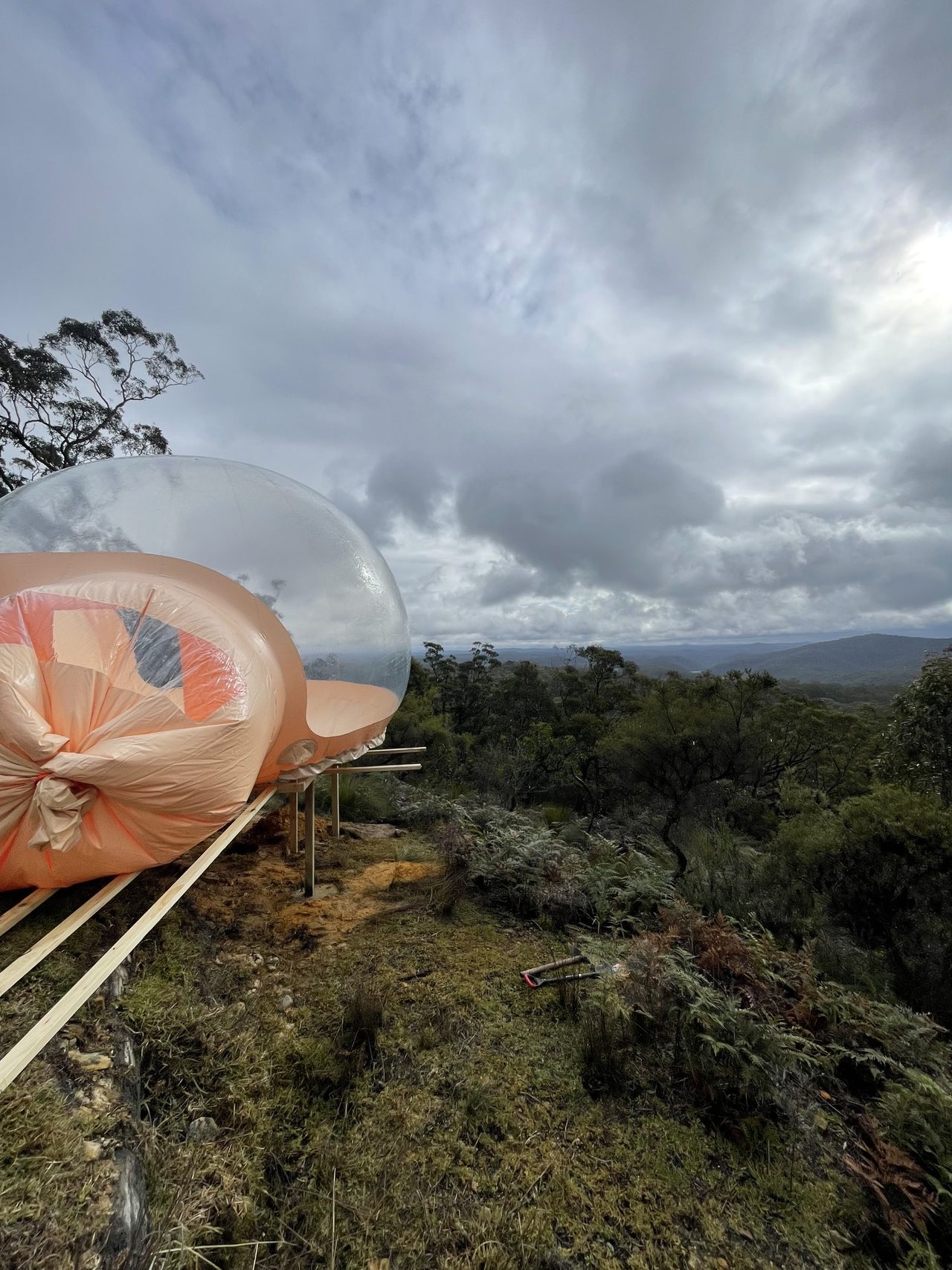 A transparent geodesic dome part-inflated on the finished timber deck, still partly in its wrapping, under heavy cloud with bushland falling away behind it.