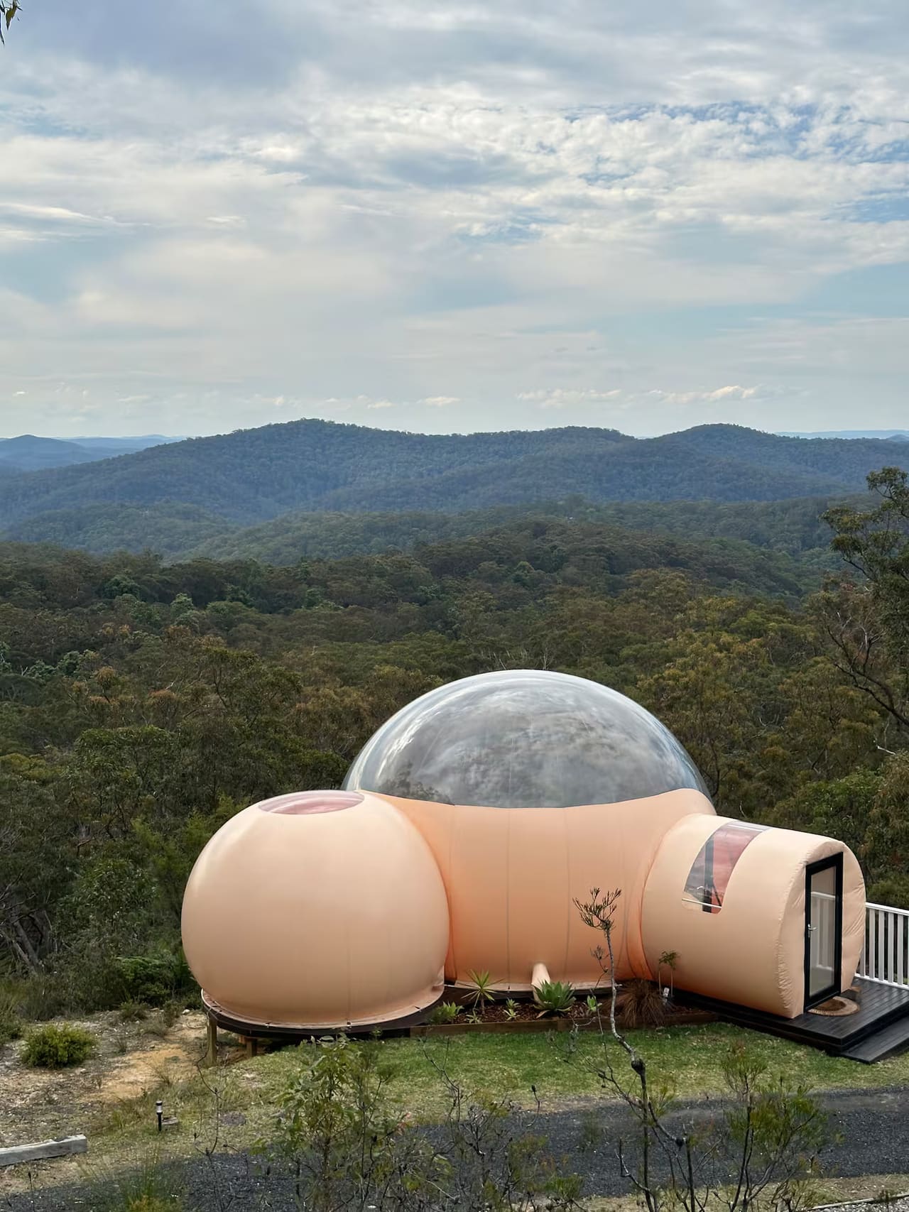 The finished bubble stay on the ridge: a peach inflatable pod with a clear dome, looking out over forested ranges under a cloudy sky.
