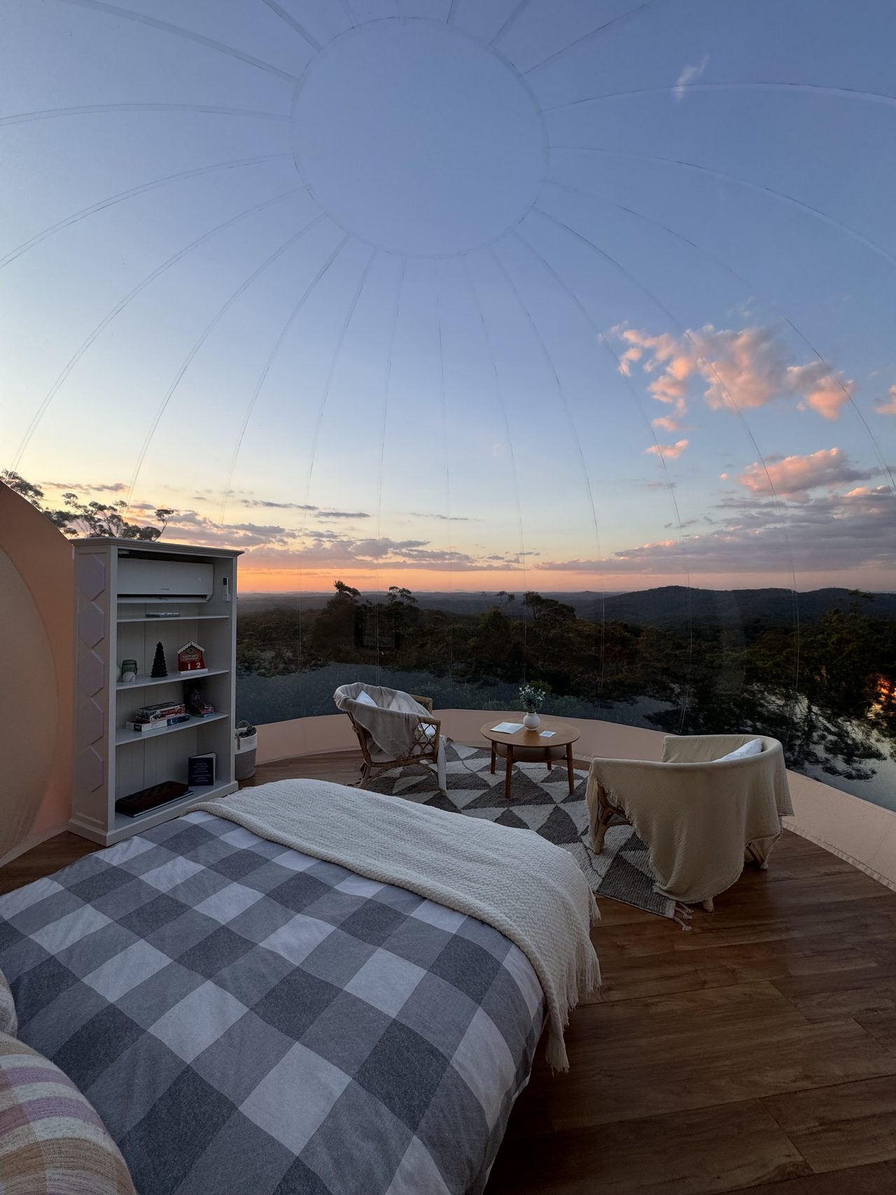 Inside the finished dome at sunset: a made bed with checked bedding, two armchairs and a side table, looking out through the clear wall to ranges and a pink and gold sky.