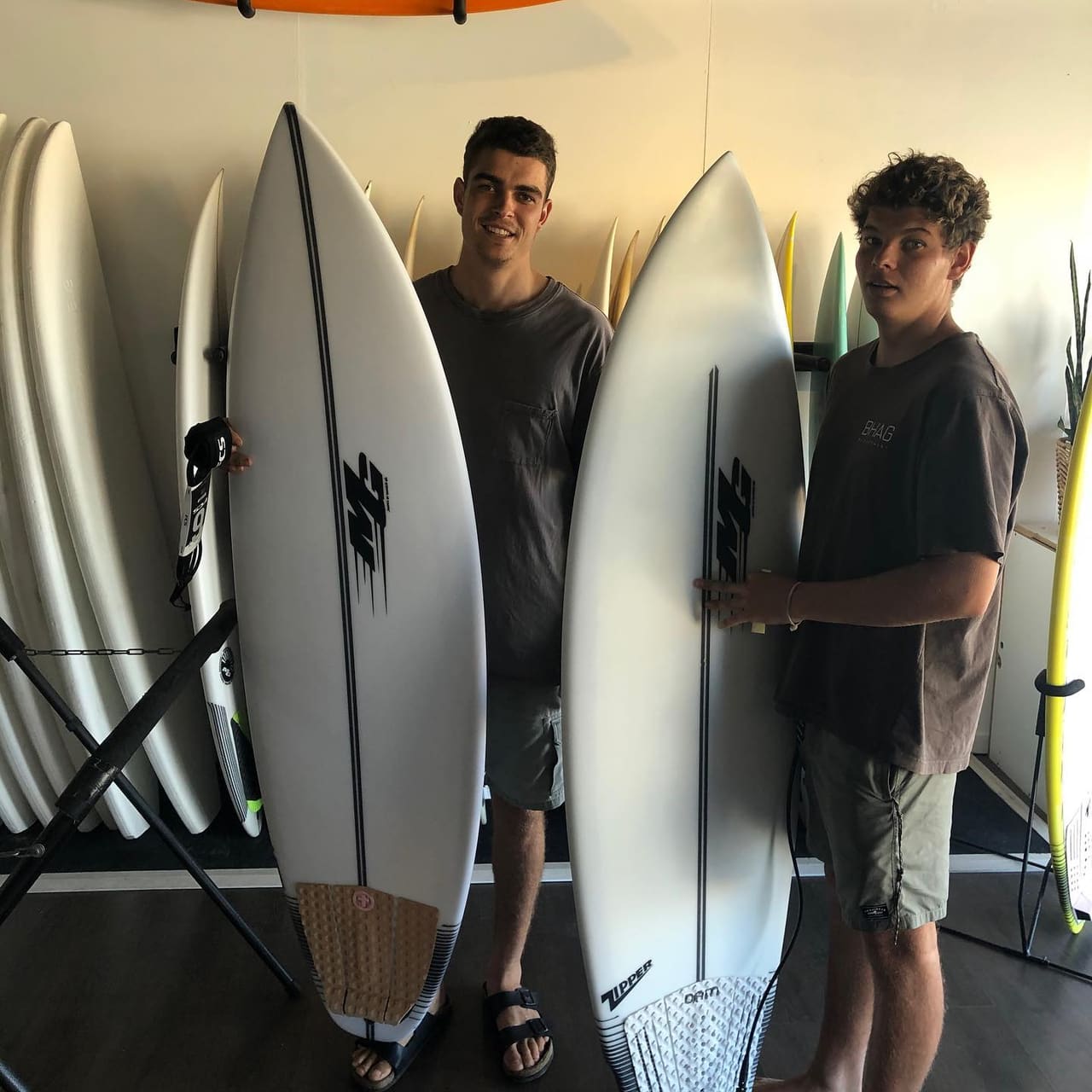 Two young men in a surf shop, each holding a brand-new white surfboard, with racks of boards on the wall behind them.